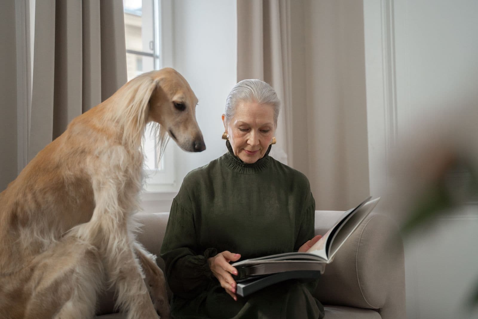 A grey-haired woman on a sofa reading a large book while a long-haired sighthound sits beside her and looks down at the page